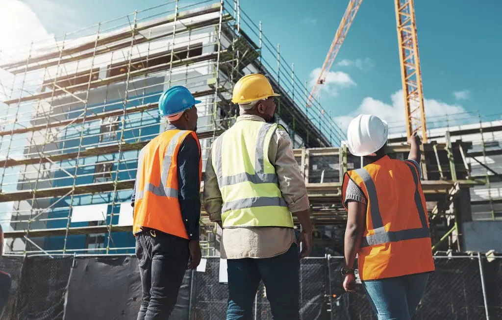 Three construction workers in safety gear observe a building site with scaffolding and a crane under a blue sky.