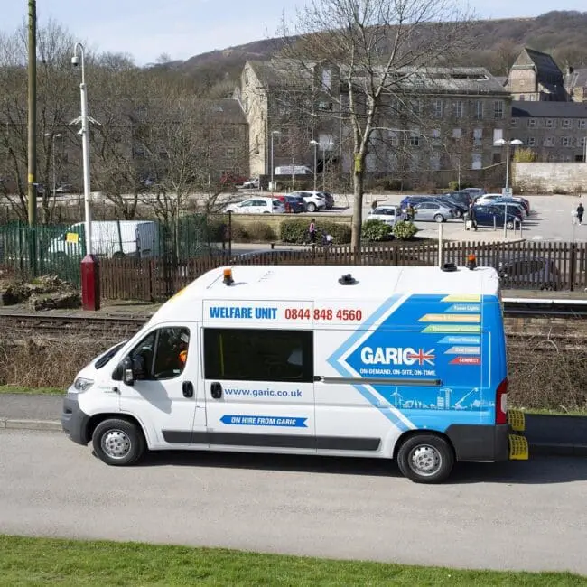 A white Garic welfare van parked near train tracks with buildings and trees in the background.