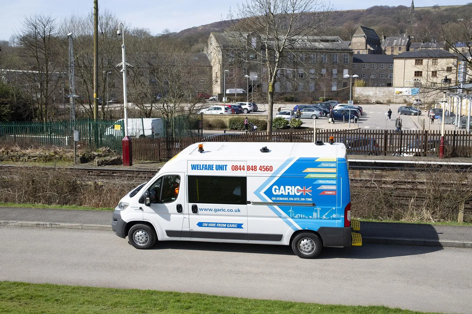 A white Garic welfare van parked near train tracks with buildings and trees in the background.