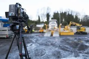 A video camera on a tripod films construction vehicles and workers at a muddy outdoor site.