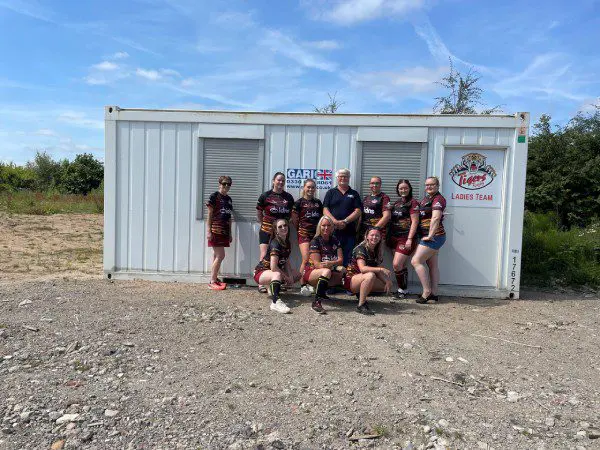 Eight women in sports uniforms pose together in front of a white storage container on a sunny day.