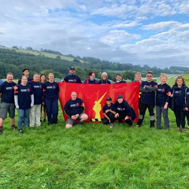 A group of people in matching shirts pose on grass with a red flag at a campsite with tents and hills.