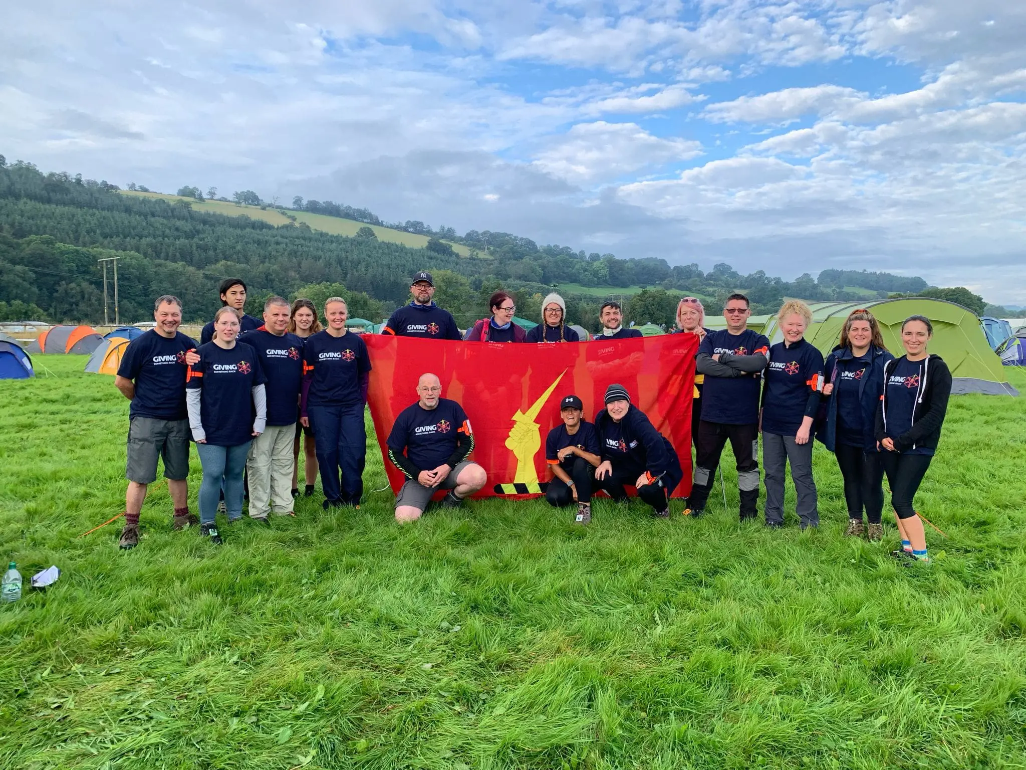 A group of people in matching shirts pose on grass with a red flag at a campsite with tents and hills.