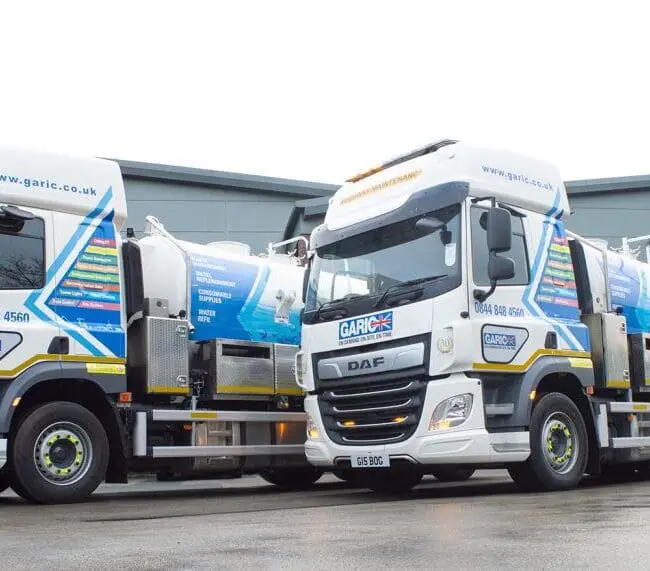 Two blue and white Garic vacuum tanker trucks parked outside a modern industrial building.
