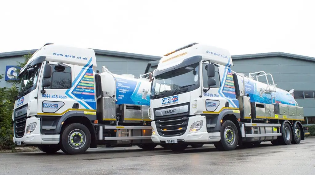 Two blue and white Garic vacuum tanker trucks parked outside a modern industrial building.