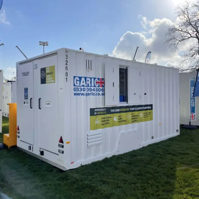 White portable toilet cabin with UK flags, contact info, and eco-friendly labels, set up outdoors on grass.