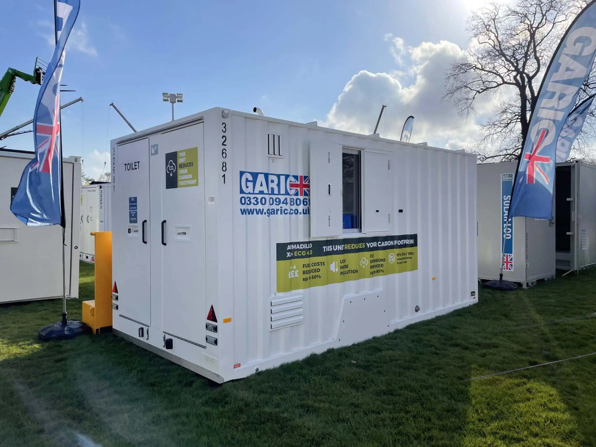 White portable toilet cabin with UK flags, contact info, and eco-friendly labels, set up outdoors on grass.