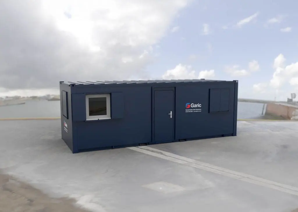 The Accommodation 24, a dark blue portable cabin with a door and two windows, is positioned on a concrete surface beneath a cloudy sky.