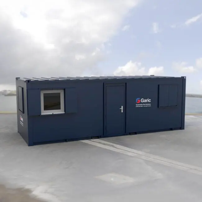 The Accommodation 24, a dark blue portable cabin with a door and two windows, is positioned on a concrete surface beneath a cloudy sky.