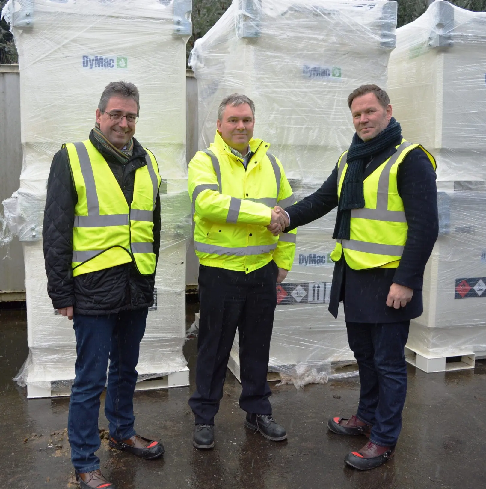 Three men in high-visibility jackets shake hands in front of large wrapped industrial equipment outdoors.