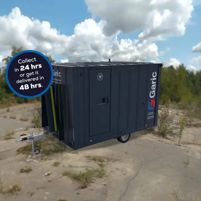 The Armadillo 12, a dark portable cabin trailer, is set on a rough concrete area with trees and clouds in the background.
