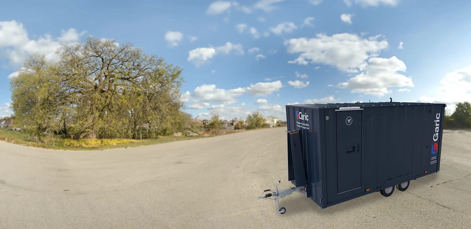 The Armadillo 18, a dark blue mobile trailer, is parked on an empty paved area with trees and a partly cloudy sky in the background.