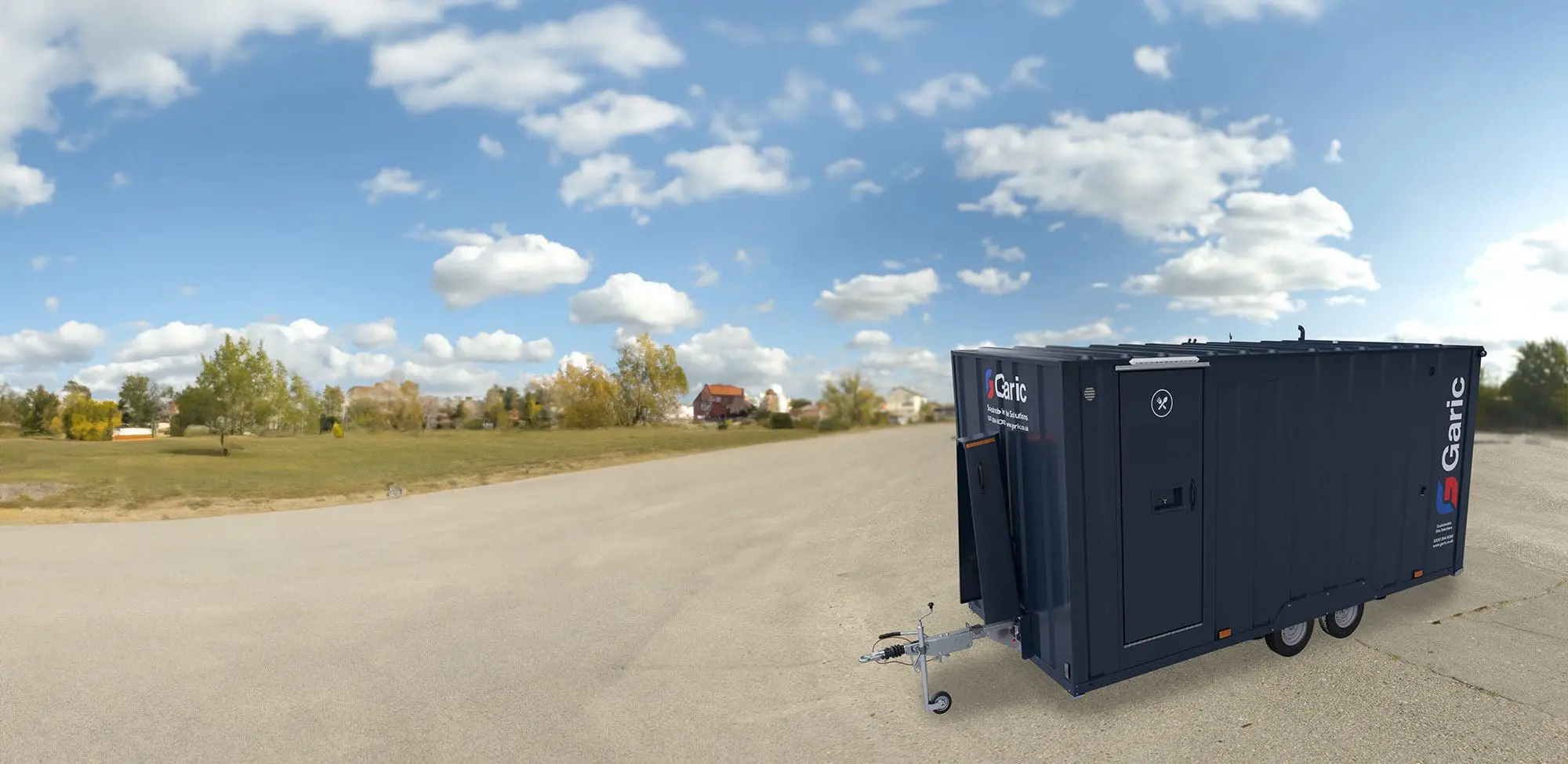 The Armadillo 18 For Hire, a dark mobile trailer, is parked on pavement surrounded by green grass and trees beneath a partly cloudy sky.