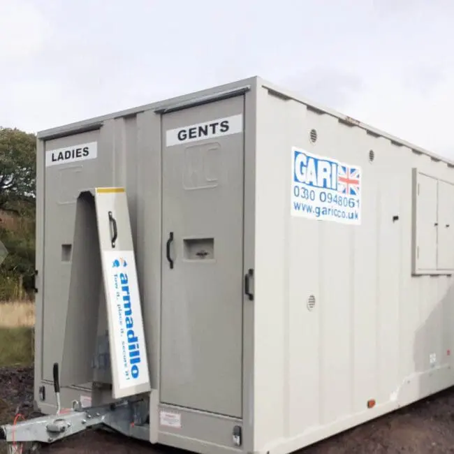 A portable toilet trailer with separate ladies and gents entrances, parked on a grassy area outdoors.
