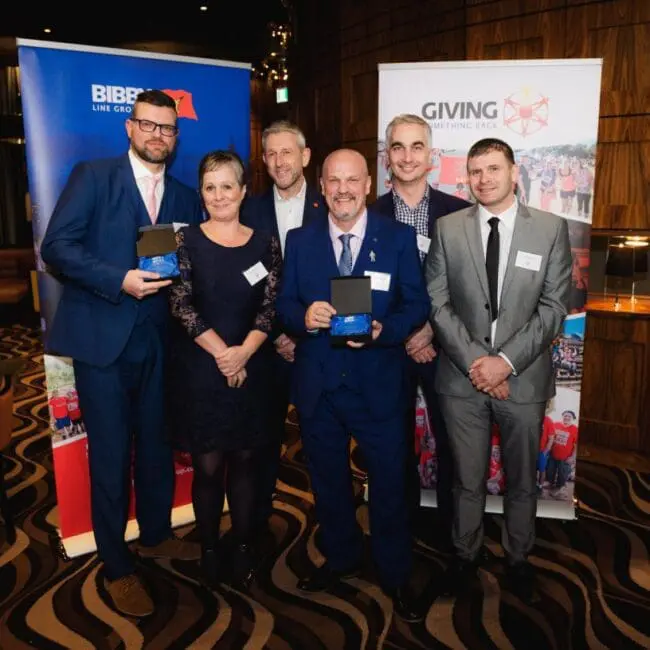 Six people in business attire pose together at an awards event, holding plaques and smiling at the camera.
