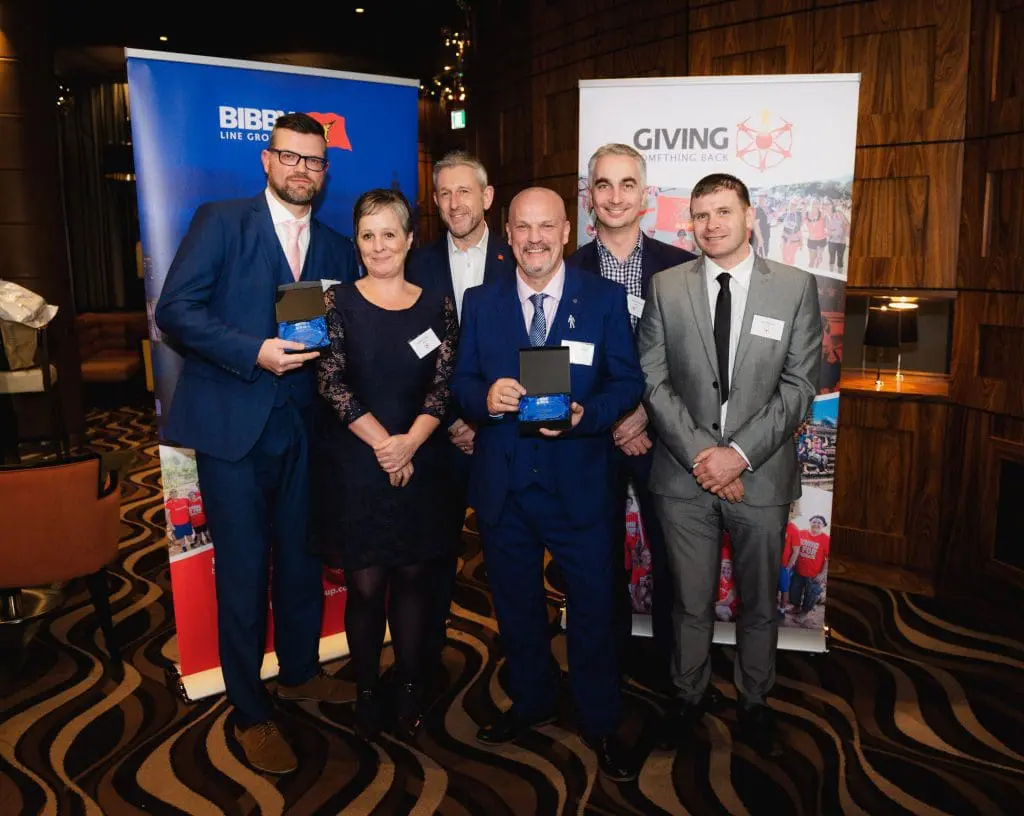 Six people in business attire pose together at an awards event, holding plaques and smiling at the camera.