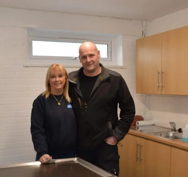 A woman and a man stand together in a small, tidy room with white walls and wooden cabinets.