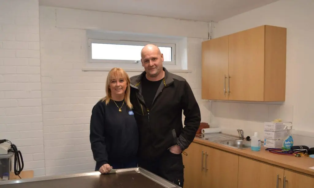 A woman and a man stand together in a small, tidy room with white walls and wooden cabinets.