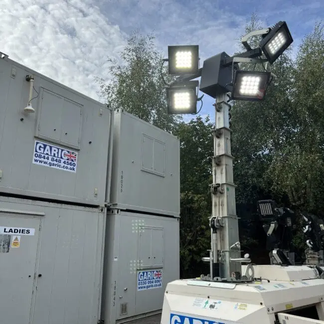 A tall portable floodlight stands near portable cabins and trees under a partly cloudy sky.
