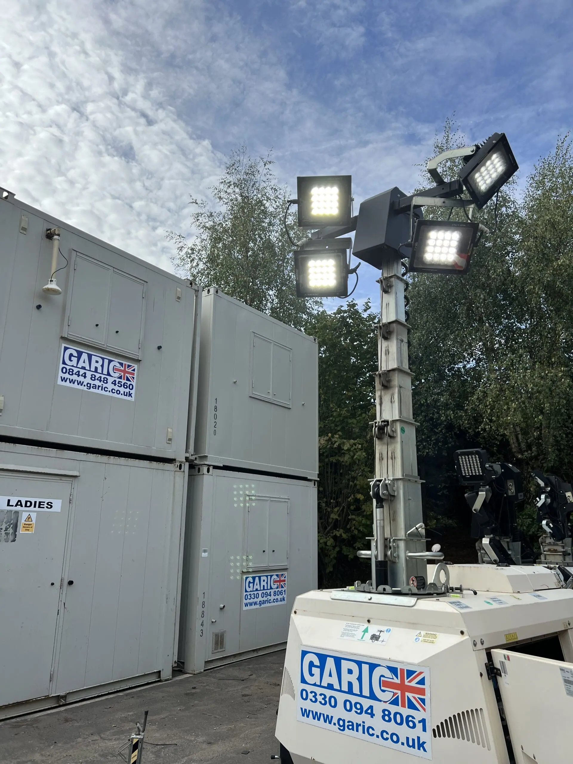 A tall portable floodlight stands near portable cabins and trees under a partly cloudy sky.