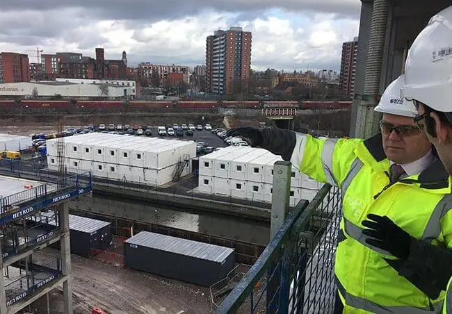 Two men in hard hats and hi-vis jackets discuss a construction site overlooking city buildings and portable offices.