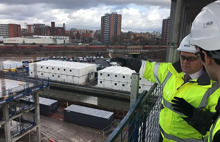 Two men in hard hats and hi-vis jackets discuss a construction site overlooking city buildings and portable offices.