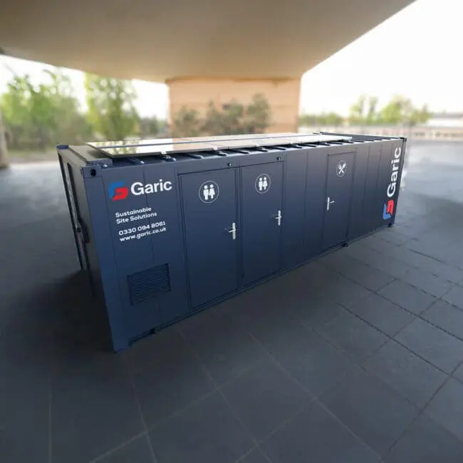 A blue Combi Cabin 24 Eco portable restroom unit sits outdoors on a tiled surface under a large structure.