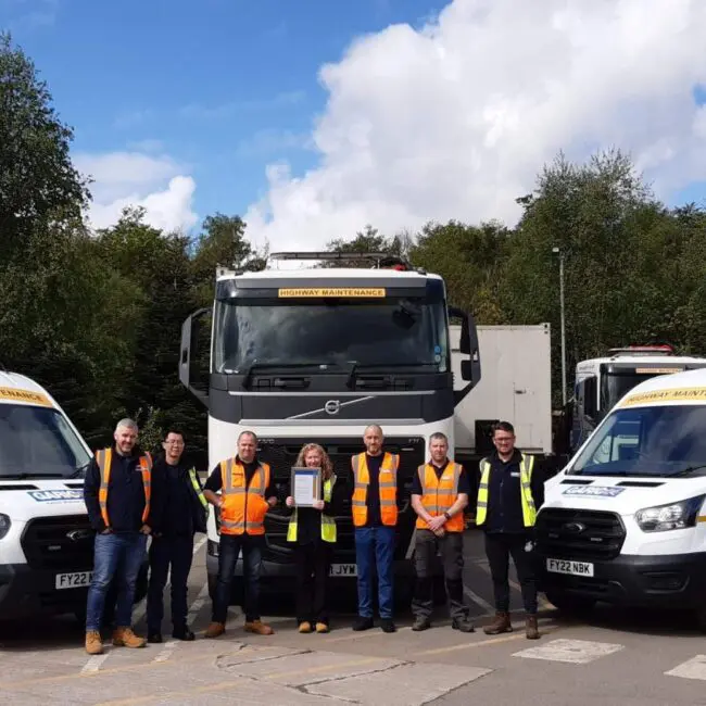 Eight people in safety vests stand in front of two vans and a truck in a parking lot on a sunny day.
