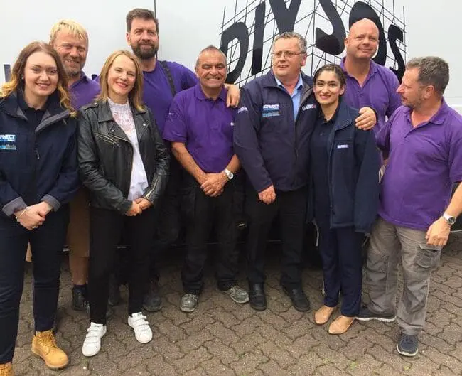 A group of nine people, smiling, stand in front of a van with the DIY SOS logo.