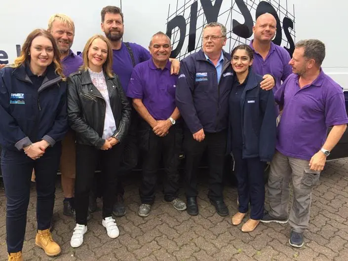 A group of nine people, smiling, stand in front of a van with the DIY SOS logo.