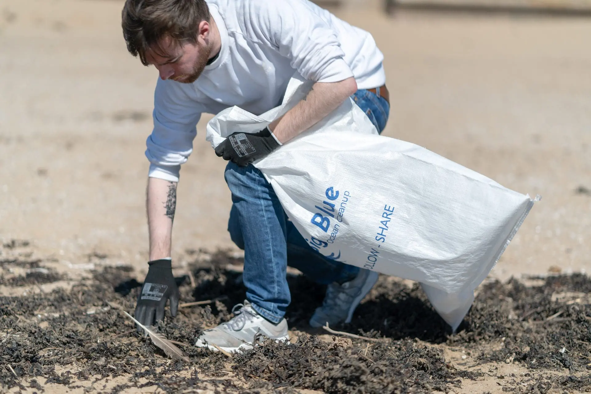 Man wearing gloves picking up trash on a beach and putting it into a large white bag.