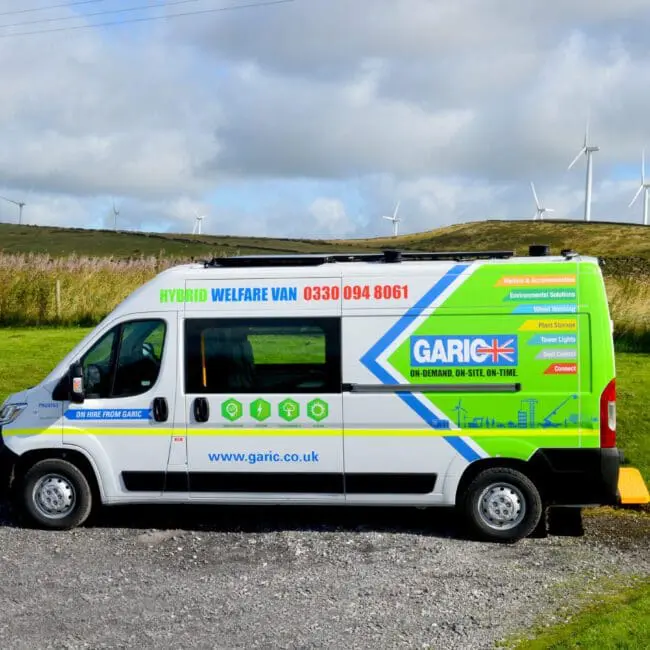 A brightly colored Garic hybrid welfare van parked on gravel with wind turbines in the background.