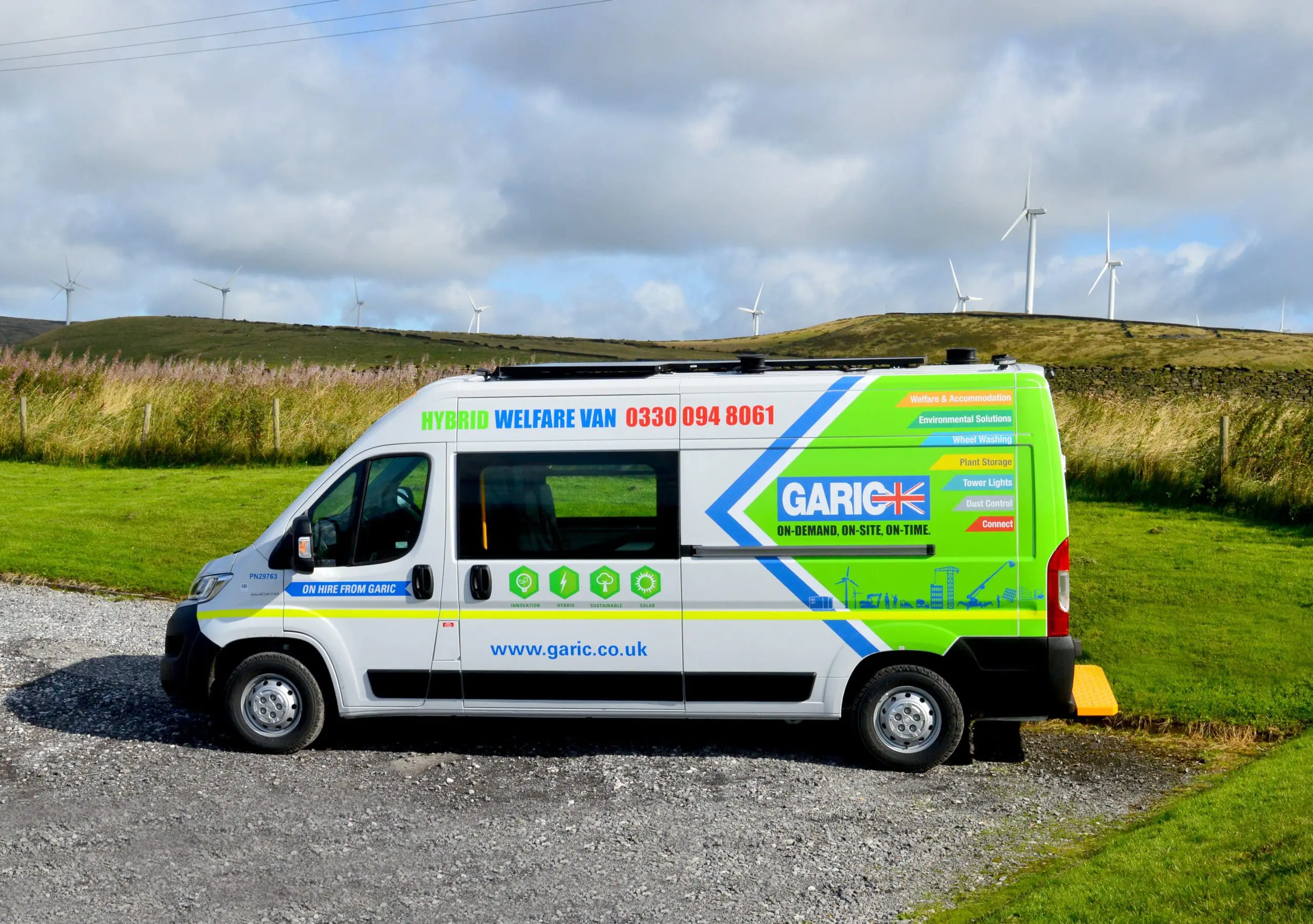 A brightly colored Garic hybrid welfare van parked on gravel with wind turbines in the background.