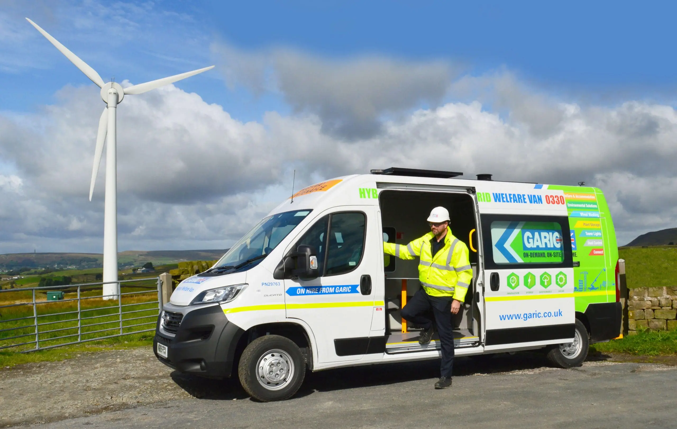 A worker in a hi-vis jacket stands by an open van door near a wind turbine in a rural area.