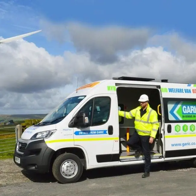 A worker in a safety vest exits a company van near a wind turbine in a rural landscape.
