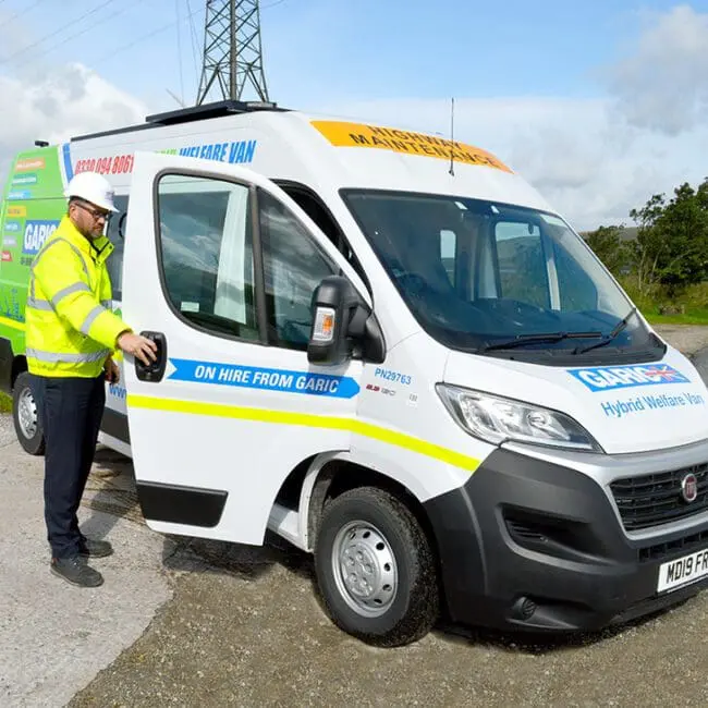 Man in high-visibility jacket and helmet opening door of a white welfare van parked on a rural road.