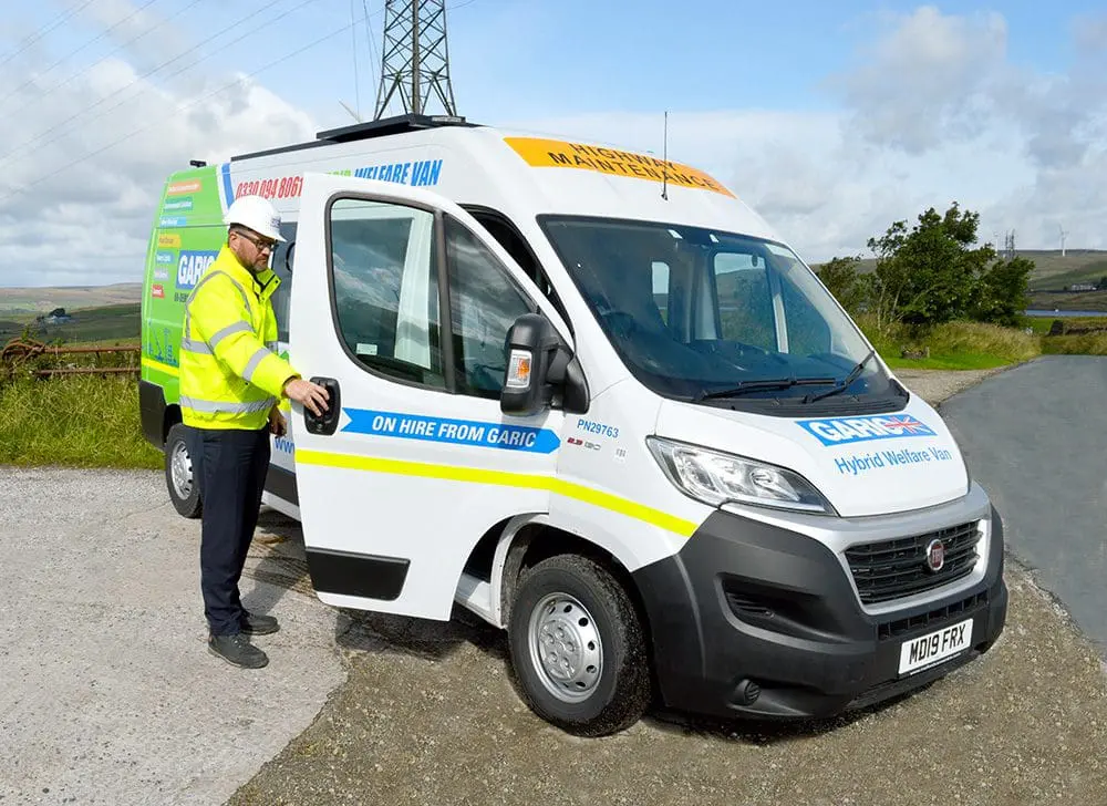 Man in high-visibility jacket and helmet opening door of a white welfare van parked on a rural road.