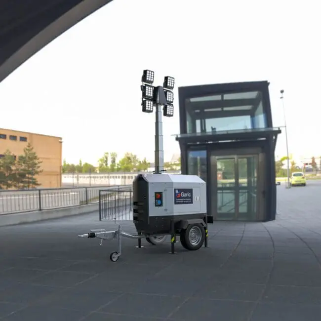 The Eco Tower Light, equipped with four large lights, is positioned on a paved outdoor area next to a modern glass building.
