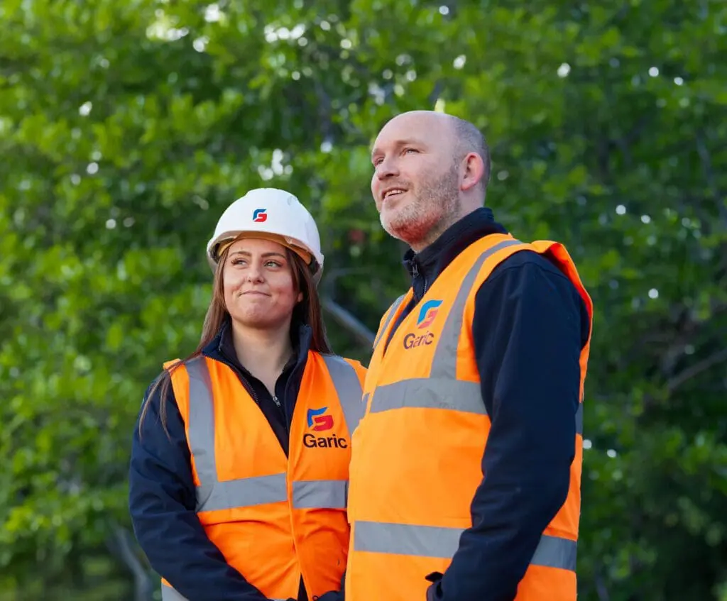 Two construction workers in orange Garic vests and hard hats stand outdoors by trees, looking up as they install a Solar Power Frame.