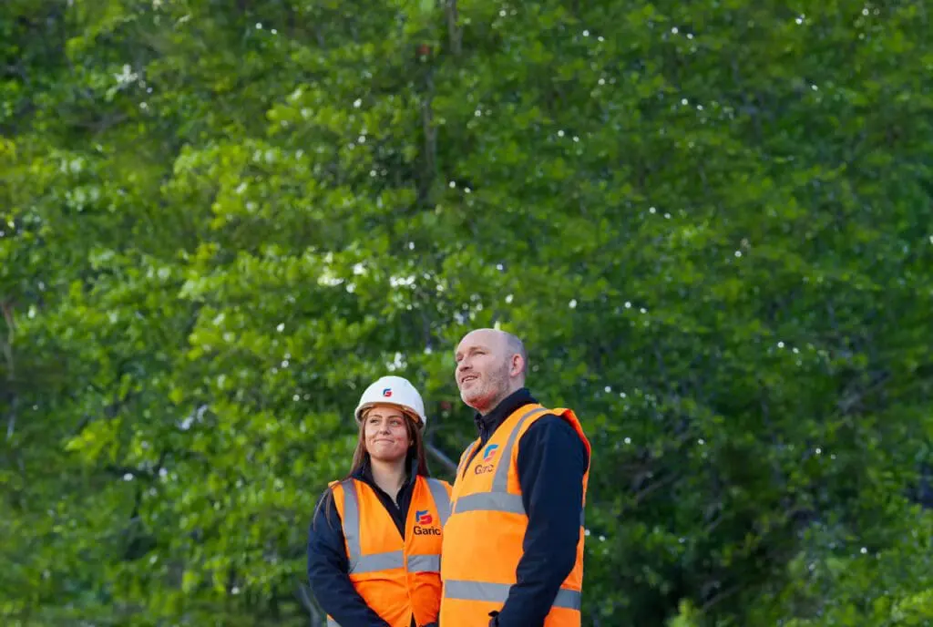 Two construction workers in orange vests stand outdoors before green trees, looking ahead. They are equipped with the Armadillo 12 Eco.