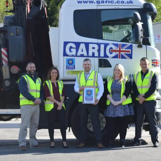 Five people in yellow safety vests stand in front of a Garic truck, holding a framed certificate.