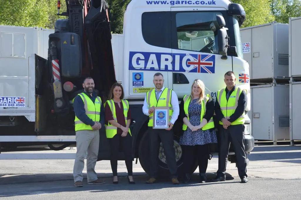 Five people in yellow safety vests stand in front of a Garic truck, holding a framed certificate.