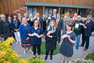 A group of people posing outdoors, several holding cards and smiling at the camera in a garden setting.