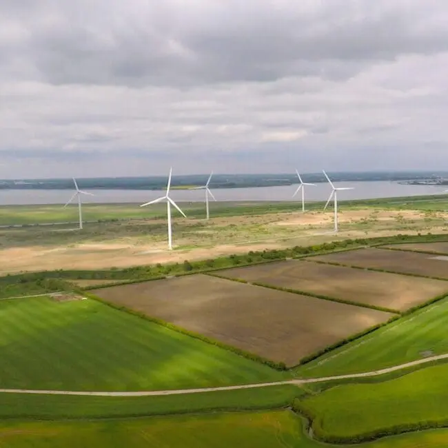 Aerial view of wind turbines on green fields near a body of water under a cloudy sky.