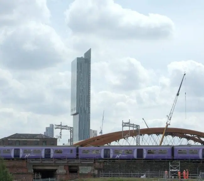 A purple train passes on a bridge with a tall modern building and cranes in the background under a cloudy sky.