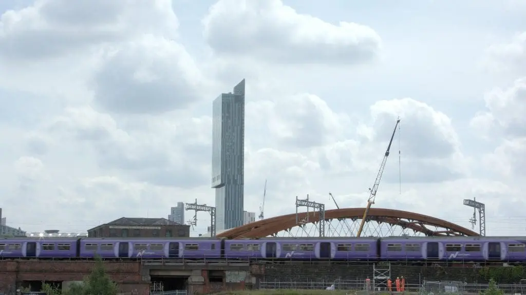A purple train passes on a bridge with a tall modern building and cranes in the background under a cloudy sky.