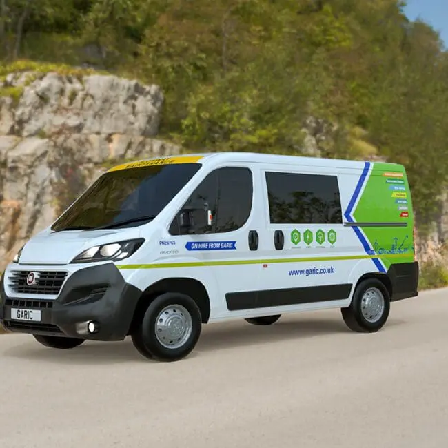 The Welfare Van Eco, a white electric van with colorful graphics, drives along a rural road bordered by rocky hills and green trees.