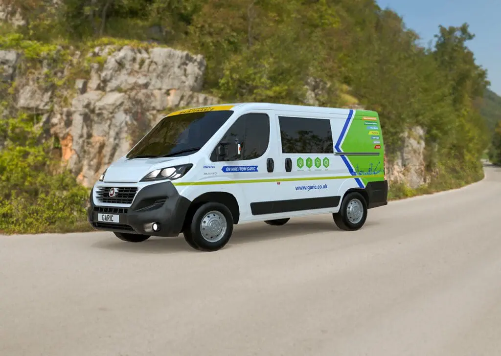 A white and green Welfare Van Eco electric service van drives on a winding road beside a rocky hillside and trees.