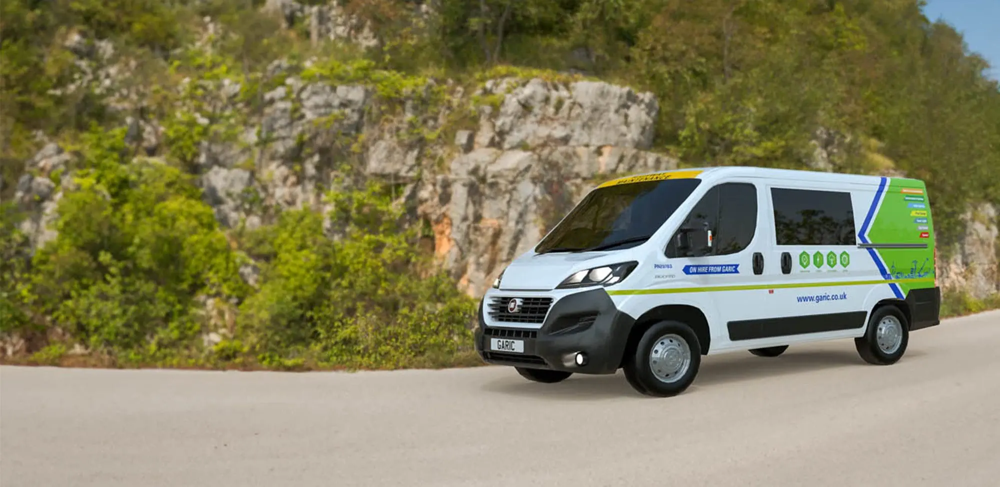 A Welfare Van Eco, featuring green and blue detailing, drives along a road next to a rocky hillside covered with trees.