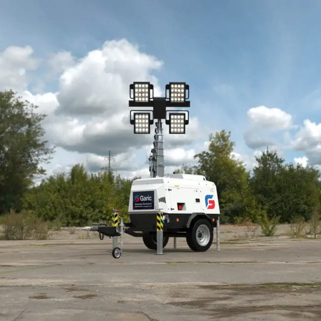 A Hybrid Tower Light with four large floodlights stands on an empty paved area with trees in the background.